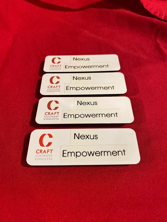 Four white name badges with two lines of printed text and a small printed mark on the left, laid out in a row on red fabric