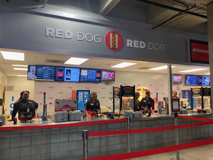People standing behind a long service counter indoors, with lit menu screens mounted above it, stacked trays and drink taps along the counter, and red-belted crowd barriers on poles across the foreground