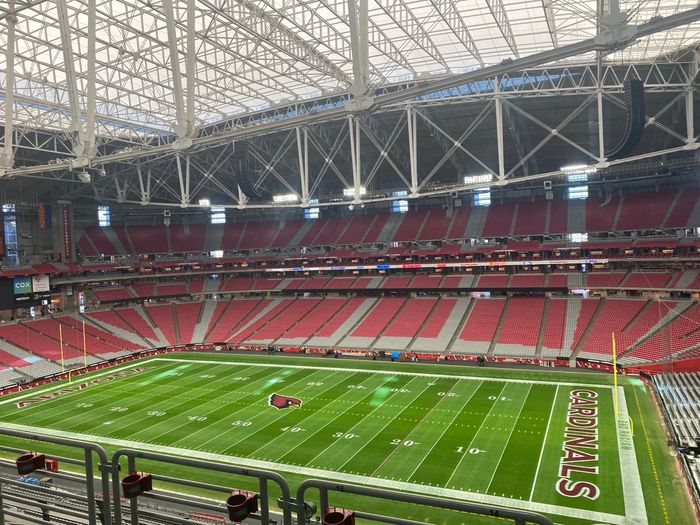 An empty green playing field seen from high in the stands, with tiers of red and grey seating curving around it, roof beams overhead and goalposts at either end