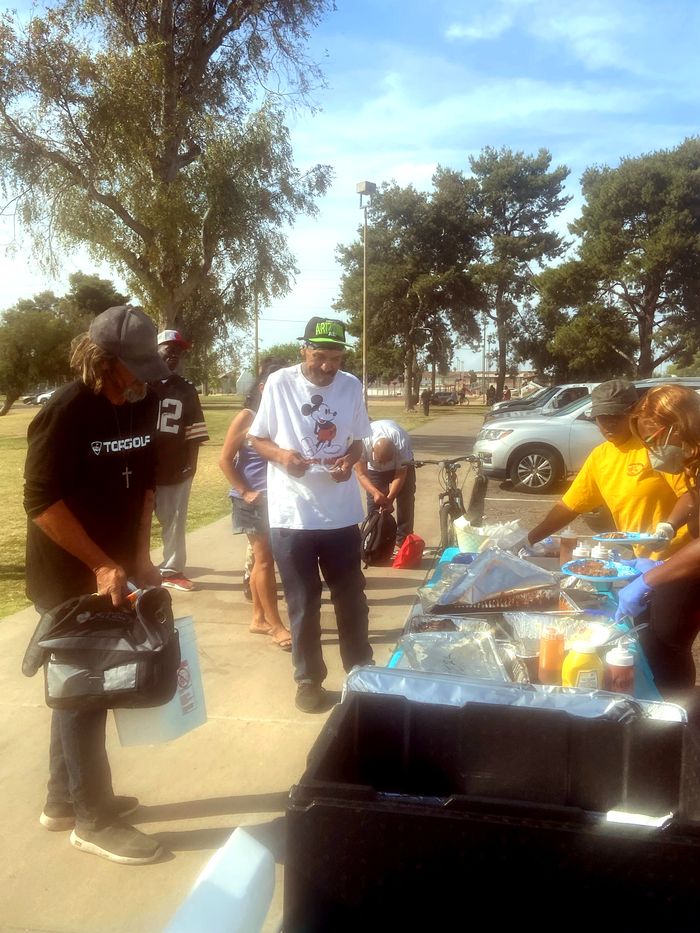 People gathered at tables outdoors in a park, taking food from foil trays, with someone serving from one side and others standing nearby holding bags and plates