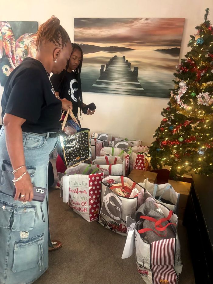 Two people standing among gift bags and boxes gathered on the floor indoors beside a decorated tree, with framed artwork on the wall behind them